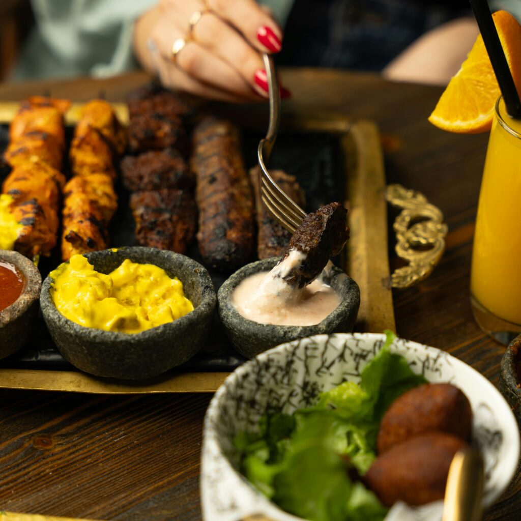 Close-up of a hand holding a fork with grilled meat over a variety of sauces, an appetizing meal setting.