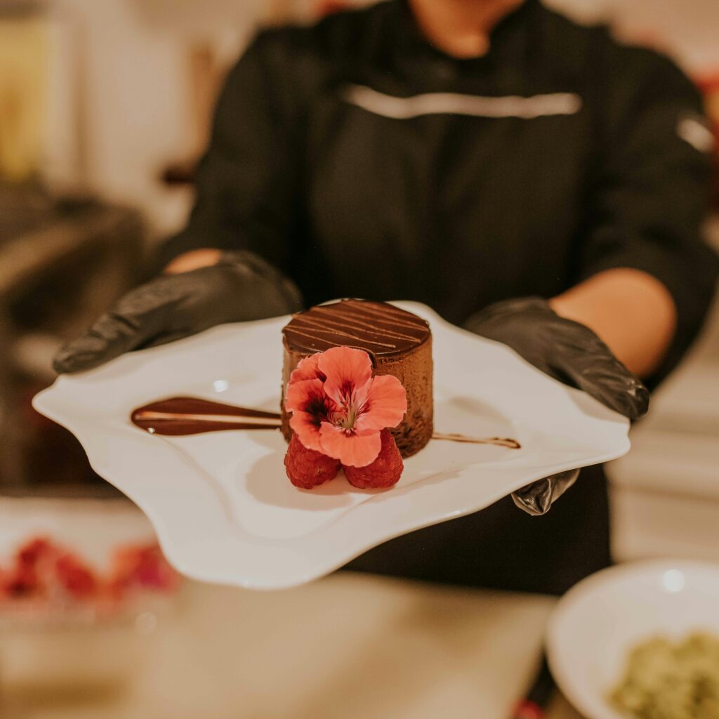 Close-up of a chocolate dessert garnished with raspberries and a flower on a ceramic plate, held by a chef in gloves.